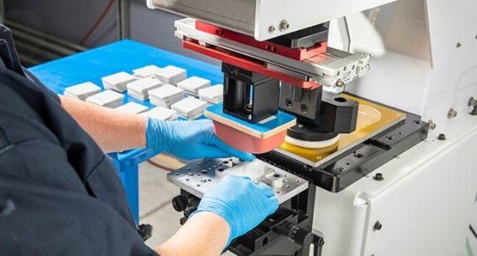 Operator aligning a custom part under a pad printing machine silicone pad in a precision manufacturing facility