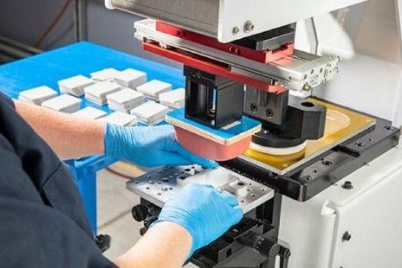 Operator aligning a custom part under a pad printing machine silicone pad in a precision manufacturing facility